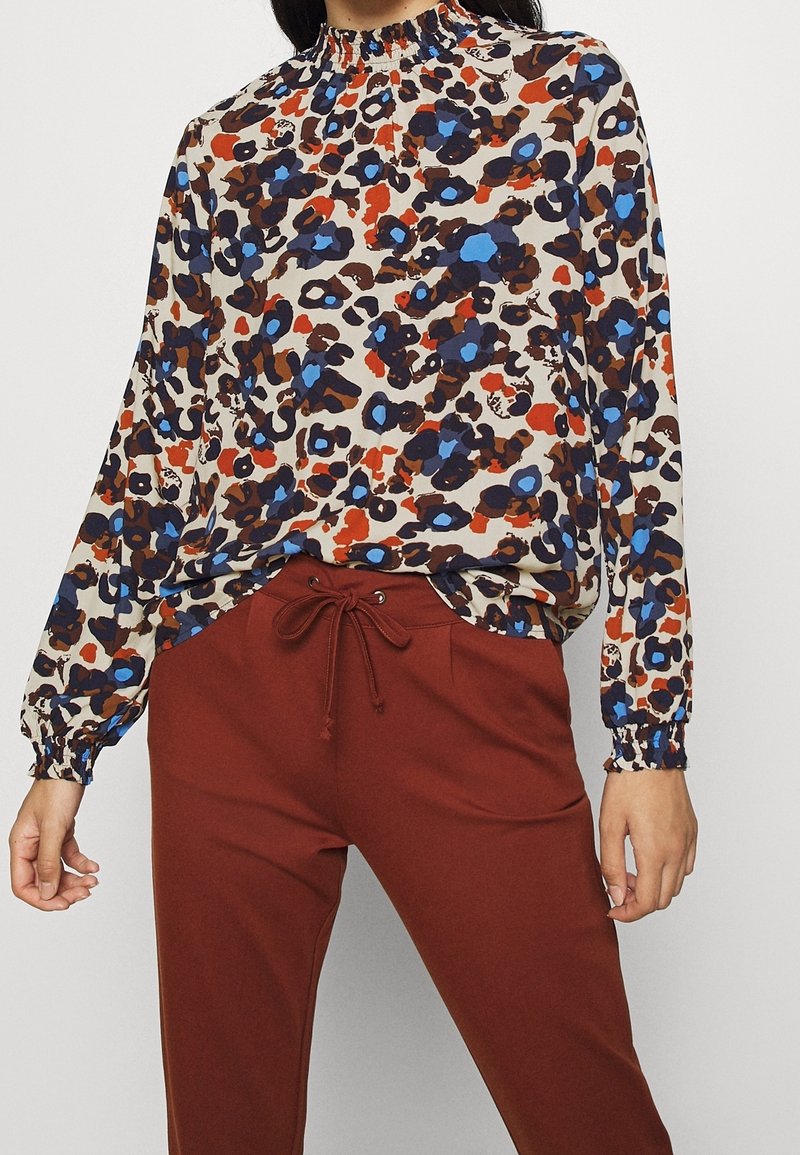 Blůza v světle béžové barvě s multikolorovaným leopardím potiskem; má nařasený výstřih a dlouhé rukávy, doplněná o kalhoty v rezavé barvě.