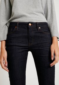 Jeans de mezclilla azul oscuro con costura naranja, diseño de cinco bolsillos y cierre de botón metálico. Combinados con una blusa gris texturizada de manga larga.