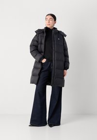 Lang svart boblejakke med hette, med quiltet tekstur og glidelås foran. Kombinert med mørkeblå vide jeans.