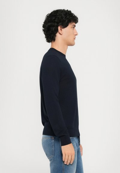 Marineblaues Rundhals-Sweatshirt aus glattem Strickmaterial, mit langen Ärmeln und einem schmalen Saum. Das Model trägt es zu hellblauen Jeans.