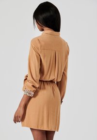 Robe à manches courtes en tissu beige, avec un col, une taille élastique et des poignets à motifs floraux pour embellir. Texture lisse et coupe décontractée.