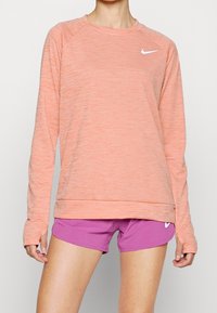 Ljus korallfärgad långärmad träningstop med ribbad textur och rund hals, kombinerad med enfärgade lila shorts som har en liten vit logotyp.