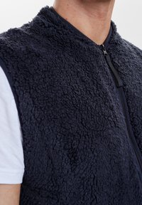 Marinblå fleeceväst med en texturerad, fluffig yta, dragkedja och rund halsringning, bärs över en vit skjorta.