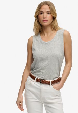 Femme aux cheveux blonds mi-longs portant un haut gris sans manches, un pantalon blanc et une ceinture tressée marron, debout avec une main dans la poche.