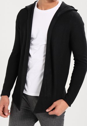 Homme portant un sweat à capuche noir ouvert sur un t-shirt blanc et un pantalon gris, debout avec une main ajustant l'ourlet du sweat à capuche.