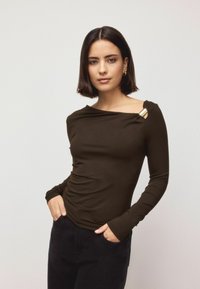 Long-sleeve, dark brown top featuring a draped neckline, gathered texture, and a gold hardware accent at the shoulder. Black pants visible.