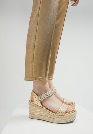 Goldene Espadrille-Sandalen mit einer geflochtenen Plattform, die glänzende Riemen und einen markierten Knöchelriemen aufweisen. Kombiniert mit goldenen, strukturierten Hosen.