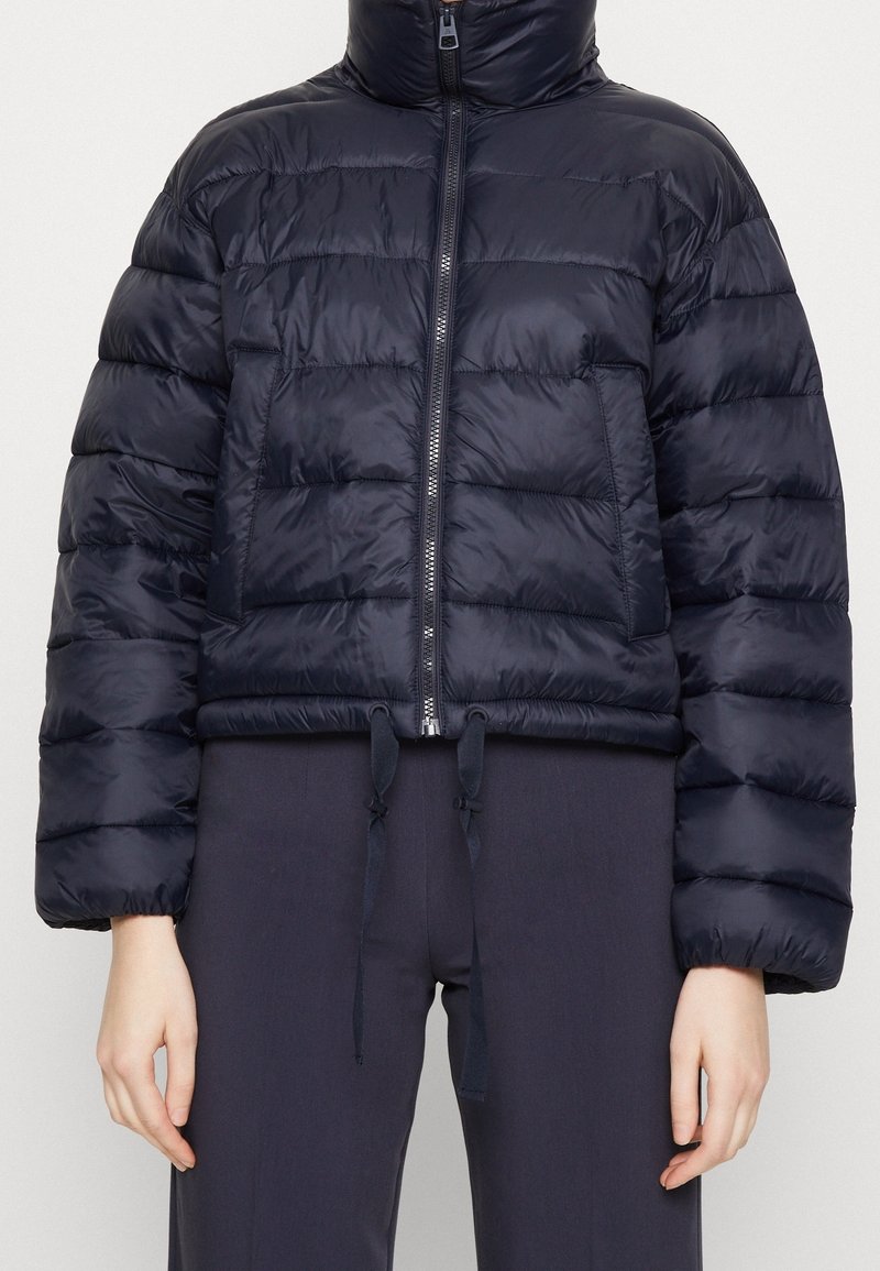 Tmavě námořnická prošívaná bunda s vysokým límcem, zapínáním na zip, nadměrným prošívaným vzorem, elastickým lemem a šňůrkami na stažení.