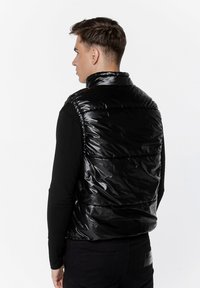 Czarna kamizelka puchowa z błyszcząca powierzchnią, wysokim kołnierzem i poziomym pikowaniem. Noszona na czarnej koszuli z długim rękawem.