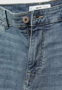 Gros plan sur un jean en denim bleu partiellement dézippé avec bouton attaché, couture visible, et étiquette de marque MNG à l'intérieur de la ceinture.