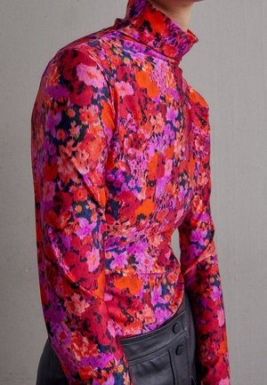 Persoon draagt een nauwsluitende coltrui met een levendig bloemmotief in rood, paars, roze en oranje, gecombineerd met zwarte, hooggetailleerde broek.