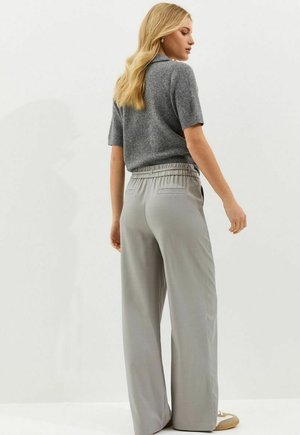 Vrouw met blond haar draagt een grijze korte mouw trui en lichtgrijze wijde broekspijpen, staand opzij tegen een effen lichte achtergrond.