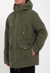 Parka aislante de color verde oliva con capucha. Presenta cierres de botón, múltiples bolsillos frontales y cordón en el dobladillo. Textura suave.