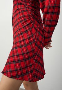 Rotes kariertes Kleid aus strukturiertem Stoff, mit langen Ärmeln, tailliertem Oberteil und ausgestelltem Saum. Seitliche Raffungen.
