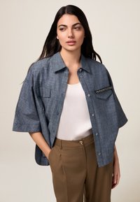 Blaues Chambray-Übergröße-Hemd mit kurzen Ärmeln, Fronttaschen und dekorativen Hardware-Details; kombiniert mit braunen, taillierten Hosen.