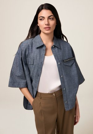 Blaues Chambray-Übergröße-Hemd mit kurzen Ärmeln, Fronttaschen und dekorativen Hardware-Details; kombiniert mit braunen, taillierten Hosen.