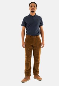 Marineblauwe polo met korte mouwen en textuurstof, gecombineerd met een bruine corduroy broek en beige schoenen, staande tegen een witte achtergrond.