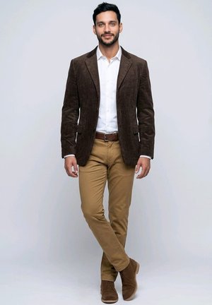 Hombre con blazer de pana marrón, camisa blanca, pantalones color arena y zapatos marrones, de pie con las piernas cruzadas frente a un fondo claro y liso.