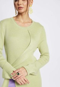Maglione a costine di colore verde chiaro con maniche lunghe, caratterizzato da un dettaglio incrociato e abbinato a un sottogonna viola.