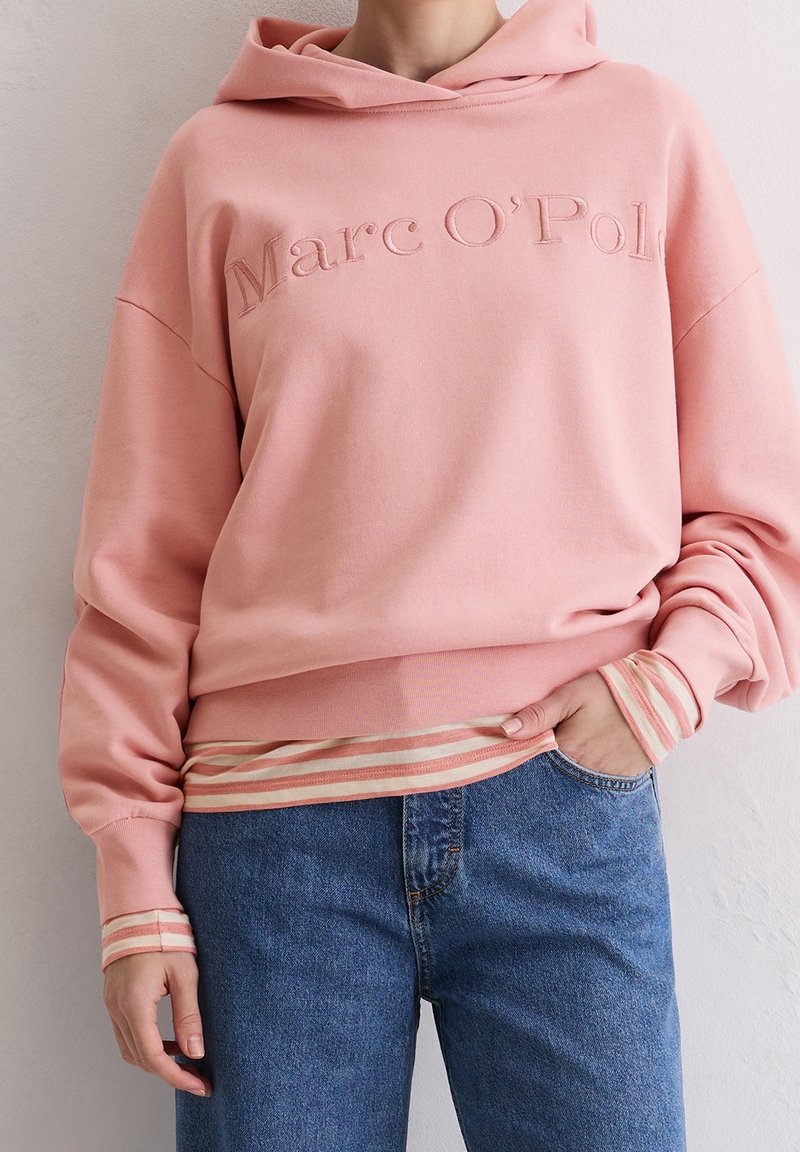 Personne portant un sweat à capuche rose Marc O’Polo par-dessus un t-shirt rayé à manches longues avec un jean bleu, une main dans la poche, contre un mur blanc.