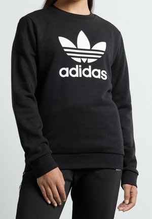 Osoba ubrana w czarną bluzę Adidas z białym logo trefoil i napisem, w połączeniu z czarnymi spodniami, na tle jednolitego jasnego tła.