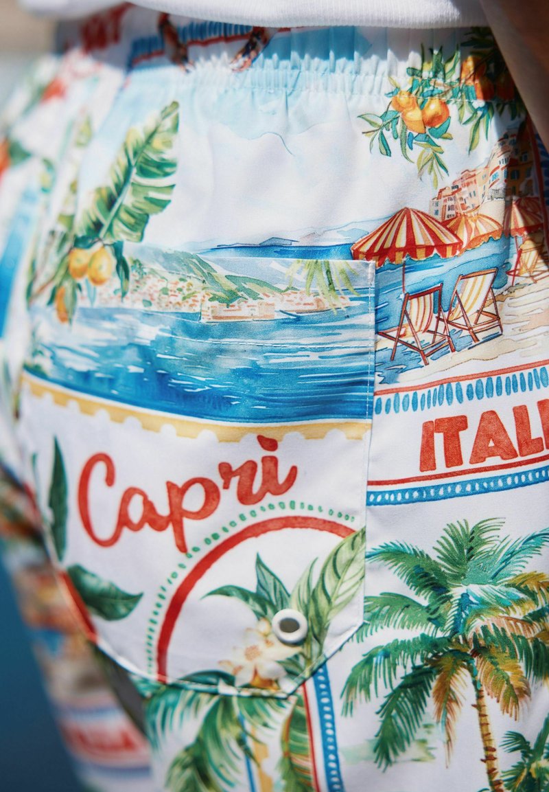 Uszónadrág, ami élénk grafikus nyomattal ábrázolja Capri-t pálmafákkal, napernyőkkel és strandfotelekkel egy könnyű, fehér anyagon.