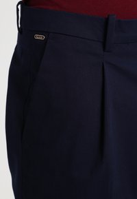 Granatowe spodnie uszyte z tkaniny mieszanej bawełnianej, cechujące się plisami, eleganckim fasonem oraz akcentem w postaci metki Scotch na pasku.