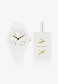 Reloj de pulsera de silicona blanca con acentos dorados, esfera minimalista con indicadores y manecillas delgadas; incluye etiqueta blanca a juego con el logo dorado.