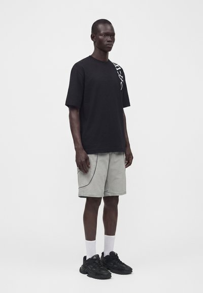 Άνδρας στέκεται φορώντας μαύρο γραφικό t-shirt, ανοιχτόγκρι σορτς με καμπυλωτές ραφές, λευκές κάλτσες και μαύρα αθλητικά παπούτσια σε μονόχρωμο φόντο.