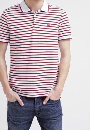 Homme portant un polo blanc à rayures horizontales bordeaux et un jean bleu, debout les mains dans les poches contre un fond blanc.