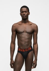 Άνδρας μοντέλο με σκούρο δέρμα, φορώντας μαύρα εσώρουχα με κόκκινη ζώνη μέσης με το λογότυπο "Calvin Klein", στέκεται μπροστά σε απλό φωτεινό φόντο.