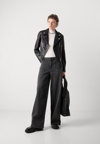 Zwarte leren motorjacket over een witte hoogsluitende top, gecombineerd met wijde donkere grijze jeans en een zwarte totebag. Chunky zwarte laarzen.