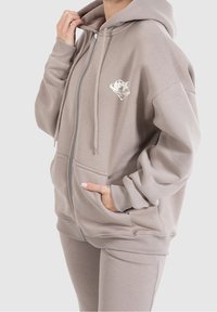 Felpa con zip color khaki realizzata in morbido tessuto, con maniche lunghe, cappuccio regolabile con lacci e tasche frontali. Presenta un piccolo logo grafico sul petto.