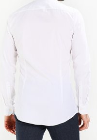 Biała koszula z długim rękawem, prostym kołnierzem, mankietami na guziki i zaokrąglonym dołem, wykonana z gładkiej tkaniny. Bez widocznych wzorów ani ozdób.