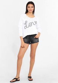 Vit stickad tröja med en grå "Love"-design, kombinerad med svarta jeansshorts och svarta flip-flops. Modellen står mot en neutral bakgrund.