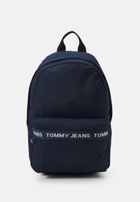Granatowy plecak z tkaniny o zaokrąglonym kształcie, z białą taśmą z logo "TOMMY JEANS" oraz górnym uchwytem. Prosty design i miękka faktura.
