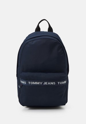 Mochila de tela azul marino con forma redondeada, que presenta una banda con el logo blanco "TOMMY JEANS" y un asa de transporte en la parte superior. Diseño simple y textura suave.