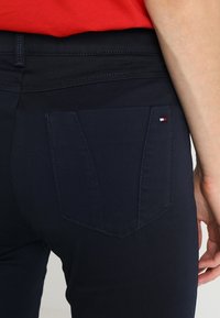 Marineblauwe broek met een achterzak met een driehoekig ontwerp en een klein rood-wit logo-accent. Gladde stofstructuur en pasvorm.