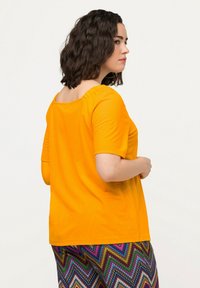 Camiseta amarilla de manga corta con escote redondo y detalle fruncido en la parte trasera, combinada con pantalones de chevron multicolores con un patrón.