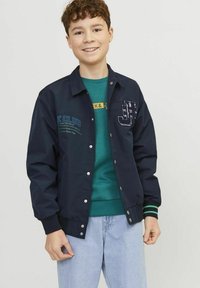 Giacca blu navy con bottoni a pressione, con testo ricamato e un logo. Il colletto e i polsini hanno dettagli a righe verdi. Indossata sopra un maglione verde.