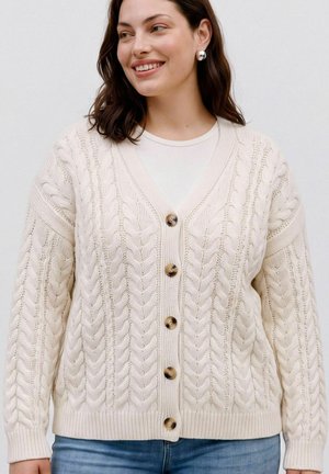 Femme portant un cardigan en maille torsadée crème avec de gros boutons sur un haut blanc, associé à un jean bleu, souriante et regardant sur le côté.