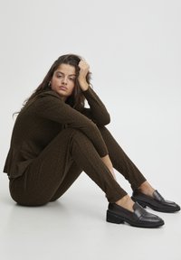 Brown patterned knitted outfit with a fitted top and tapered pants, paired with black loafers. Model sits on the floor, resting her chin on her hand.