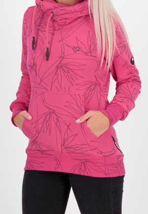 Vrouw draagt een roze hoodie met zwarte lijnpatronen van bladeren, een hartspeld op de borst, handen in de voorkant van de zak, gecombineerd met zwarte broek.