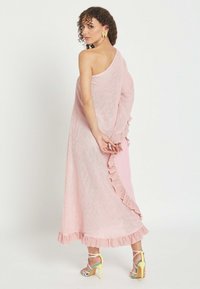 Mujer con cabello rizado que lleva un vestido rosa pálido de un solo hombro con bordes con volantes y sandalias de tacón alto multicolor, de pie con las manos detrás de la espalda.
