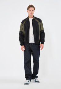 Svart og olivengrønn vindjakke med ståkrage, elastiske mansjetter og glidelås foran, kombinert med mørke denimjeans.