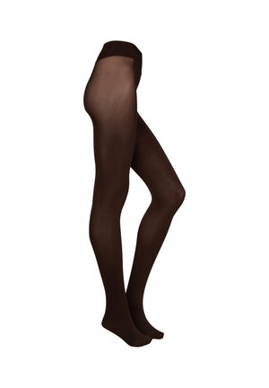 Frau trägt braunen BH und schwarze transparente Strumpfhosen, steht mit verschränkten Händen da und schaut leicht zur Seite vor einfachem Hintergrund.
