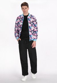 Geprinte bomberjack met een roze en blauwe bloemenpatroon, lichtblauwe afwerking, zwart T-shirt, zwarte broek en witte sneakers.