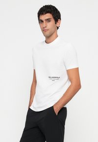 Ung mann med kort mørkt hår iført en hvit t-skjorte med kort erme med "KARL LAGERFELD"-tekst og svarte bukser, hendene i lommene.