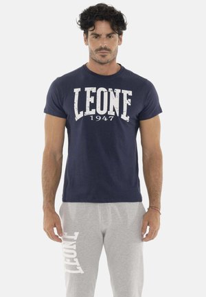 Homme aux cheveux bouclés foncés portant un t-shirt bleu marine avec le texte "LEONE 1947" et un pantalon de jogging gris clair avec le texte blanc "LEONE" sur la jambe gauche.