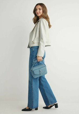 Kobieta w jasnoszarej kurtce i niebieskich dżinsach, trzymająca małą niebieską torebkę, stojąca na tle białego tła.